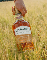 A hand holds a bottle of Aberargie Golden Promise single malt Scotch whisky standing in a field of golden barley.